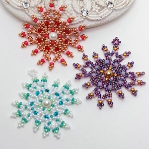 Three handcrafted, intricate beaded snowflake pendants or ornaments featuring designs in purple/gold, white/turquoise, and red/copper color palettes, displayed on an ornate white surface.
