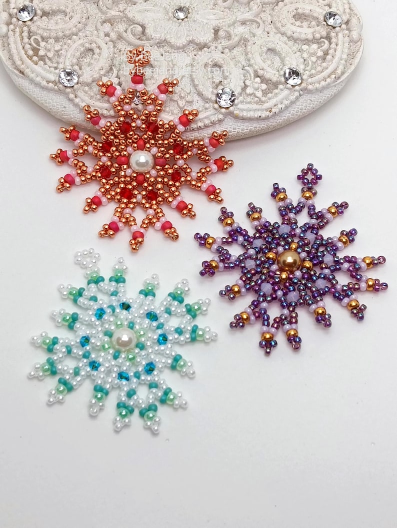 Three handcrafted, intricate beaded snowflake pendants or ornaments featuring designs in purple/gold, white/turquoise, and red/copper color palettes, displayed on an ornate white surface.