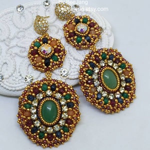 A pair of large, intricate, handcrafted drop earrings featuring an oval green aventurine center stone and extensive bead embroidery using gold, red, dark blue, green, white, and peach-colored seed beads and crystals, displayed on a white decorative surface.