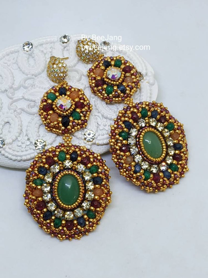A pair of large, intricate, handcrafted drop earrings featuring an oval green aventurine center stone and extensive bead embroidery using gold, red, dark blue, green, white, and peach-colored seed beads and crystals, displayed on a white decorative surface.