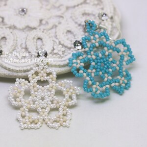 A pair of intricate, handcrafted jewelry pieces, likely earrings, featuring a snowflake or floral design made of seed beads; one is predominantly white/cream colored and the other is turquoise blue and white, displayed on a white background with lace accents.