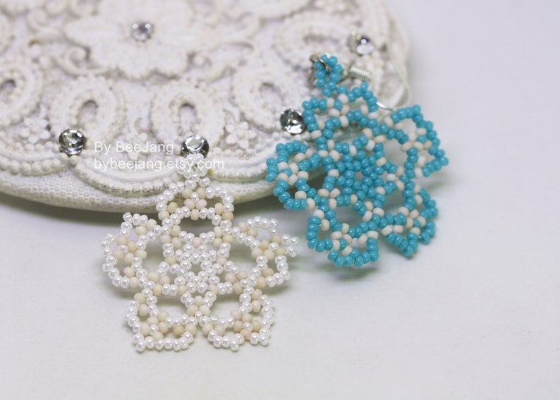 A pair of intricate, handcrafted jewelry pieces, likely earrings, featuring a snowflake or floral design made of seed beads; one is predominantly white/cream colored and the other is turquoise blue and white, displayed on a white background with lace accents.