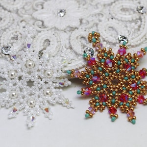 Two handcrafted, intricate beaded snowflake pendants or ornaments displayed on an ornate white lacy surface. The right snowflake features gold, red, turquoise, and pink beads, while the left snowflake features white, clear, and cream faux pearls/beads.