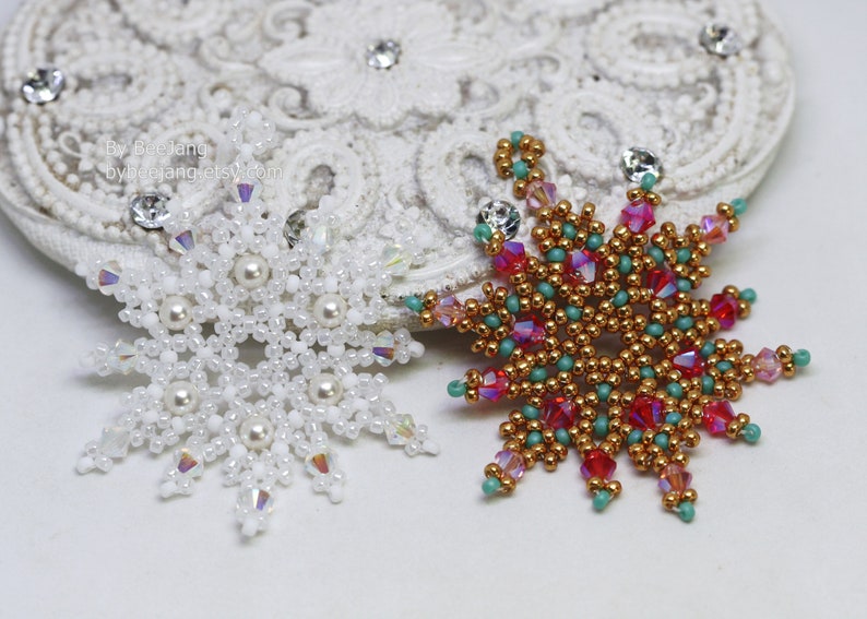 Two handcrafted, intricate beaded snowflake pendants or ornaments displayed on an ornate white lacy surface. The right snowflake features gold, red, turquoise, and pink beads, while the left snowflake features white, clear, and cream faux pearls/beads.