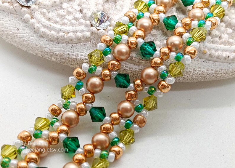 A close-up of a wide, handcrafted Right Angle Weave beaded bracelet. The intricate lattice design features emerald and olive green bicone crystals, metallic bronze round pearls, and small gold, white, and green seed beads. The beads are woven into an open, decorative pattern with a rich, textured finish.