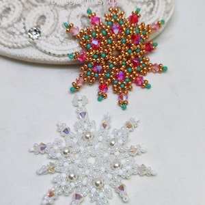 Two handcrafted, intricate beaded snowflake pendants or ornaments displayed on an ornate white lacy surface. The top snowflake features gold/copper, red, pink/magenta, and turquoise beads, while the bottom snowflake features white, clear, and cream faux pearls/beads.