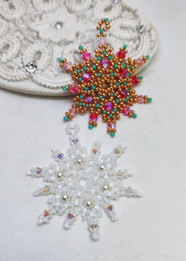 Two handcrafted, intricate beaded snowflake pendants or ornaments displayed on an ornate white lacy surface. The top snowflake features gold/copper, red, pink/magenta, and turquoise beads, while the bottom snowflake features white, clear, and cream faux pearls/beads.