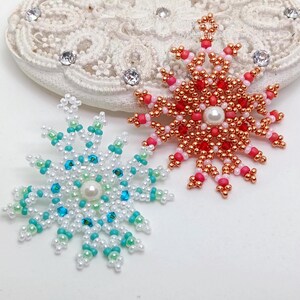 Two handcrafted, intricate beaded snowflake pendants or ornaments displayed on an ornate white lacy surface. The left snowflake features white, turquoise, and light blue beads, while the right snowflake features copper, red, and pink beads, both with central white faux pearls.