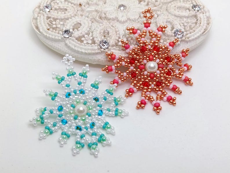 Two handcrafted, intricate beaded snowflake pendants or ornaments displayed on an ornate white lacy surface. The left snowflake features white, turquoise, and light blue beads, while the right snowflake features copper, red, and pink beads, both with central white faux pearls.