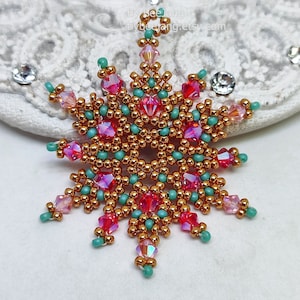A single, handcrafted, intricate beaded snowflake pendant or ornament featuring gold/copper, hot pink/magenta, and turquoise seed beads and crystal accents, displayed on an ornate white, lacy surface.