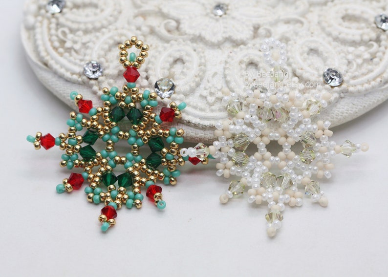 Two handcrafted, intricate beaded snowflake pendants or ornaments displayed on an ornate white surface. The left pendant features gold, turquoise, green, and red beads, while the right pendant features white, cream, and clear beads.