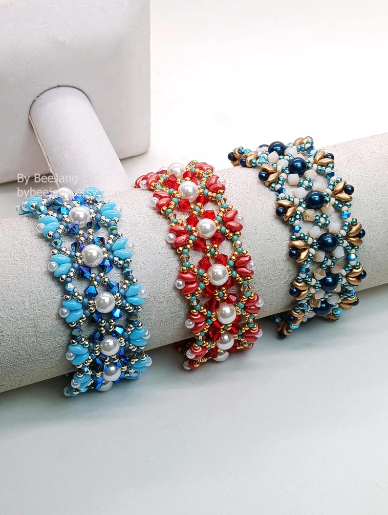 May include: Three beaded bracelets in blue, red, and gold. The bracelets feature pearls and various beads, forming a floral pattern. The bracelets are displayed on a white cylindrical form.