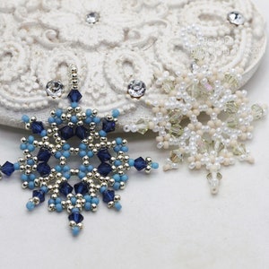 Two handcrafted, intricate beaded snowflake pendants or ornaments, one in a blue, silver, and gold color scheme, and the other in a white, cream, and clear palette, displayed on a decorative white lacy surface.
