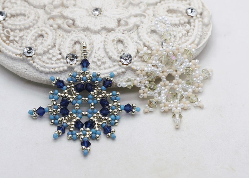 Two handcrafted, intricate beaded snowflake pendants or ornaments, one in a blue, silver, and gold color scheme, and the other in a white, cream, and clear palette, displayed on a decorative white lacy surface.