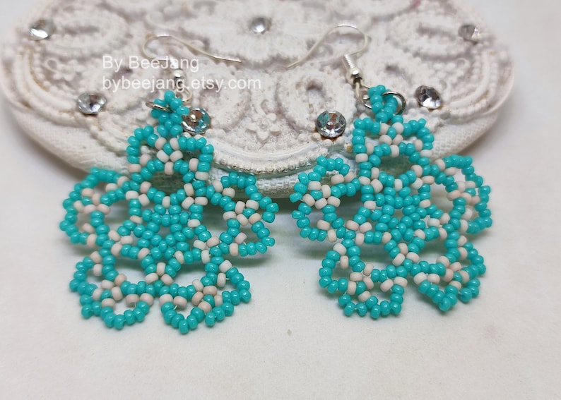 A pair of intricate, handcrafted drop earrings featuring turquoise blue and white seed beads in a snowflake or floral mandala pattern, displayed on a white lace background with small rhinestone details.