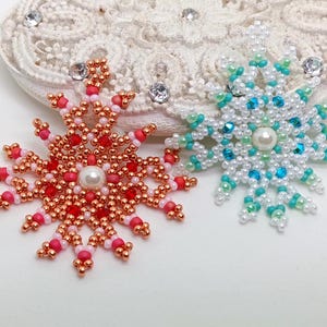 Two handcrafted, intricate beaded snowflake pendants or ornaments displayed on an ornate white lacy surface. The left snowflake features copper/rose gold, red, and white beads, while the right snowflake features white, mint green, and turquoise beads.