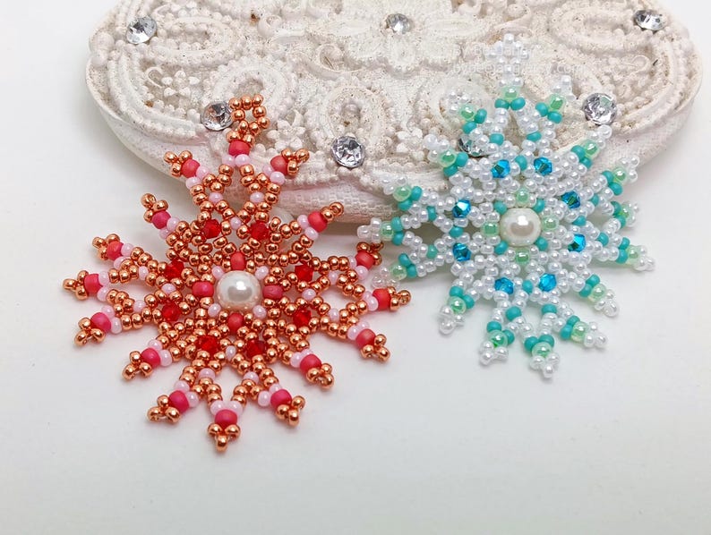 Two handcrafted, intricate beaded snowflake pendants or ornaments displayed on an ornate white lacy surface. The left snowflake features copper/rose gold, red, and white beads, while the right snowflake features white, mint green, and turquoise beads.