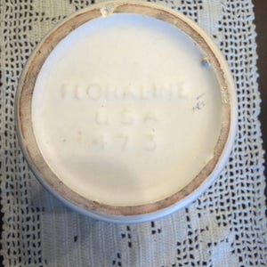 Op de afbeelding: De ronde onderkant van een witte keramische pot bevat de in reliëf aangebrachte tekst FLORALINE USA 473. De onderzijde heeft een opstaande rand met een lichtbruine, ongeglazuurde afwerking, terwijl het midden glad en wit is met fijne barstjes.
