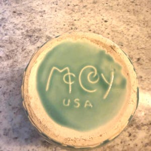 May include: The circular base of a ceramic object featuring a light teal glazed center with the embossed text "McCoy" in a stylized script font and the letters "USA" printed in a simple sans-serif typeface below the brand name.