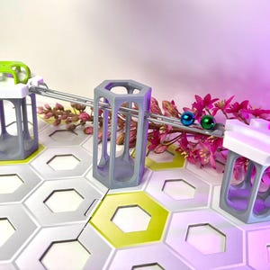 Puede incluir: Un juego de juguetes de circuito de canicas con componentes de plástico gris y blanco. El juego incluye baldosas hexagonales, estructuras de soporte y pistas. Dos canicas, una azul y otra verde, están en la pista. El fondo presenta flores rosas.
