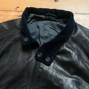 May include: Black leather jacket with a corduroy collar and a zipper closure. The jacket has two snap closures on the collar. The leather appears worn, giving it a vintage look. The jacket is a classic style.