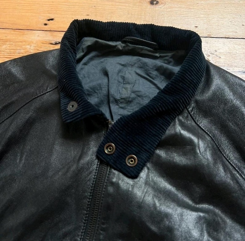 May include: Black leather jacket with a corduroy collar and a zipper closure. The jacket has two snap closures on the collar. The leather appears worn, giving it a vintage look. The jacket is a classic style.