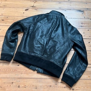 May include: Black leather jacket with a ribbed collar, cuffs, and hem. The jacket has a zipper closure and a classic bomber style. The leather appears slightly distressed, giving it a vintage look. The jacket is laid out on a wooden surface.