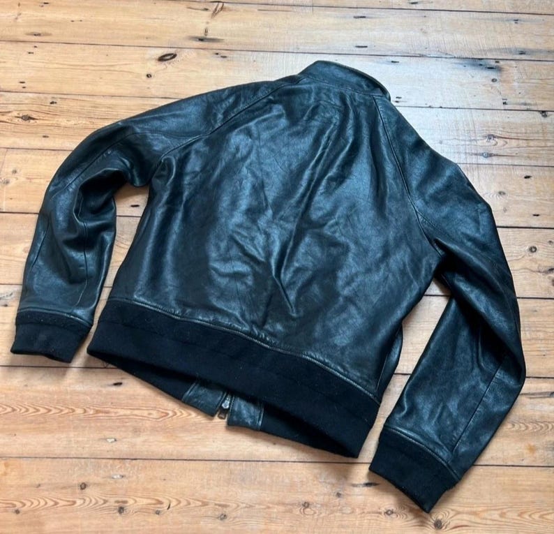 May include: Black leather jacket with a ribbed collar, cuffs, and hem. The jacket has a zipper closure and a classic bomber style. The leather appears slightly distressed, giving it a vintage look. The jacket is laid out on a wooden surface.