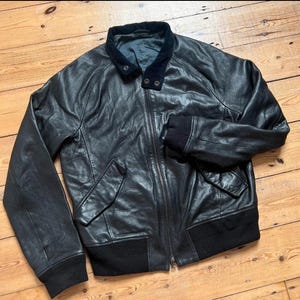 May include: Black leather bomber jacket with a zip-up front, two front pockets, and a black ribbed knit collar, cuffs, and hem. The jacket has a dark blue velvet collar and is laid out on a wooden surface.
