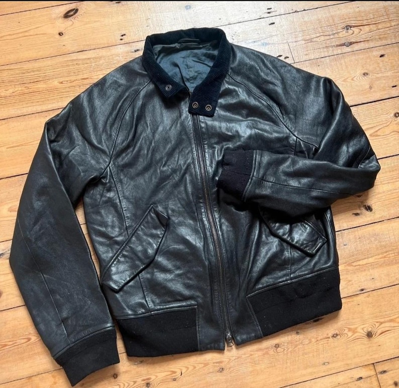 May include: Black leather bomber jacket with a zip-up front, two front pockets, and a black ribbed knit collar, cuffs, and hem. The jacket has a dark blue velvet collar and is laid out on a wooden surface.