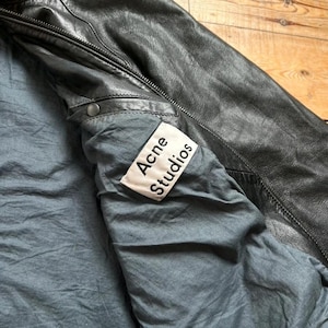 May include: Black leather jacket with a gray lining. The jacket has a zipper closure and a label that reads "Acne Studios". The jacket appears to be well-worn and has a classic design.