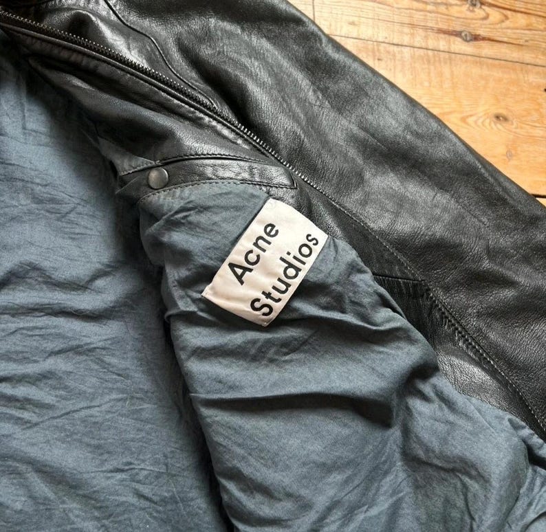 May include: Black leather jacket with a gray lining. The jacket has a zipper closure and a label that reads "Acne Studios". The jacket appears to be well-worn and has a classic design.