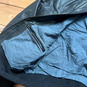 May include: Black leather jacket with a blue interior lining. The jacket features a zipper closure and a pocket with a zipper. The collar and cuffs are black. The jacket appears to be made of a soft, supple leather.
