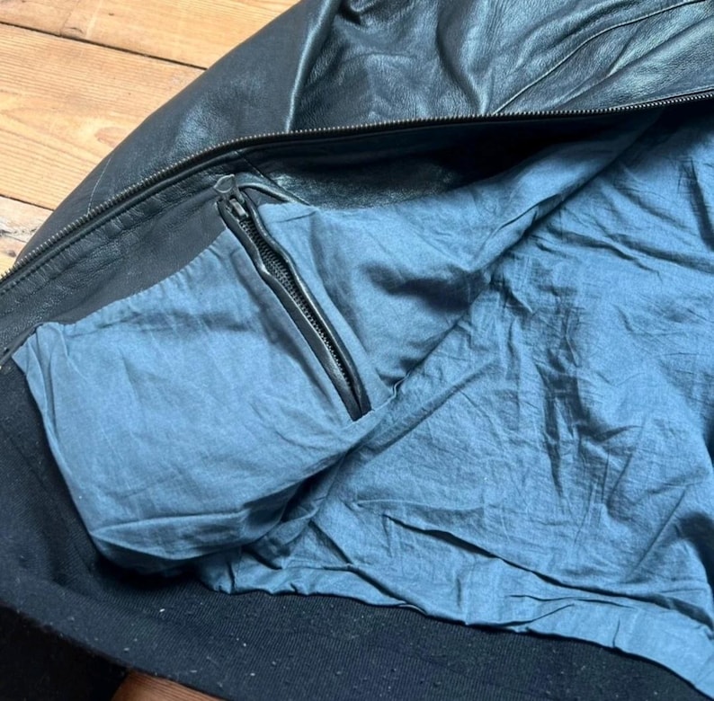 May include: Black leather jacket with a blue interior lining. The jacket features a zipper closure and a pocket with a zipper. The collar and cuffs are black. The jacket appears to be made of a soft, supple leather.