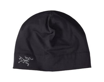 May include: A black beanie hat featuring a smooth, solid-colored fabric construction with a rounded crown. A small, light gray embroidered logo depicting a stylized fossilized creature is positioned on the lower left side of the hat.