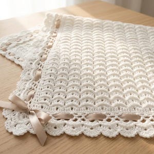 May include: A white crocheted blanket featuring a repeating shell stitch pattern and a scalloped border. A light beige satin ribbon is woven through the edge of the blanket, tied into a decorative bow at the corner of the textile piece.