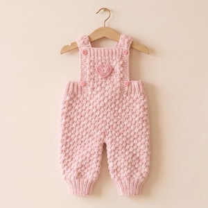 Puede incluir: Un peto de bebé de punto rosa claro con un detalle en forma de corazón en el pecho. El peto está colgado de una percha de madera. El tejido de punto tiene un patrón texturizado en forma de burbujas. El peto tiene tirantes y botones.