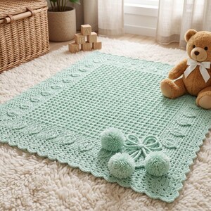 May include: A mint green crochet baby blanket with a textured pattern and scalloped edges. The blanket features decorative floral accents and pom-poms. A brown teddy bear with a white bow sits on the blanket.