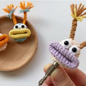 Puede incluir: Horquillas de ganchillo con forma de criaturas caprichosas. Las horquillas presentan ojos grandes, bocas abiertas y detalles decorativos en tonos naranja, amarillo, azul y morado. Las horquillas se muestran en un plato de madera y son sujetadas por una mano.