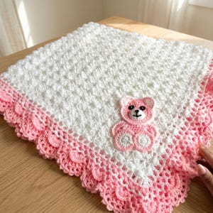 May include: A handmade, white and pink crochet baby blanket. The blanket features a textured stitch pattern and a scalloped pink border. A pink teddy bear applique is centered on the blanket, adding a charming detail. The blanket is approximately 30 inches square.