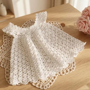 Puede incluir: Una blusa blanca de ganchillo hecha a mano con escote y mangas festoneadas, sobre un tapete de encaje color crema. La blusa presenta un delicado patrón de tejido abierto, sobre una superficie de madera clara.