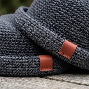 May include: Two dark gray crochet hats with a brown leather tag stitched on the brim. The hats are stacked on top of each other, showcasing the texture of the crochet and the leather detail. The hats are on a wooden surface.