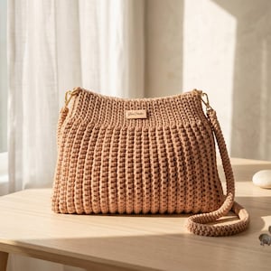 Könnte beinhalten: Eine beige gehäkelte Umhängetasche mit rechteckiger Form und einem langen, geflochtenen Riemen. Die Tasche hat ein kleines, rechteckiges Etikett mit Text. Die Tasche steht auf einem hellen Holztisch.