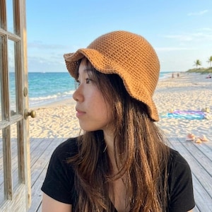 May include: A brown, crocheted bucket hat with a wavy brim. The hat is worn by a person with long brown hair, against a backdrop of a beach and ocean. The hat is a warm, neutral color.