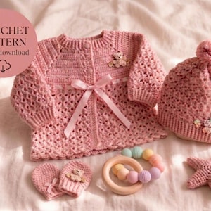 Puede incluir: Un conjunto de bebé de ganchillo rosa que incluye una chaqueta, un gorro y manoplas, con un mordedor de madera y una estrella de mar. La chaqueta tiene un lazo y adornos de osito de peluche. El gorro tiene un pompón.