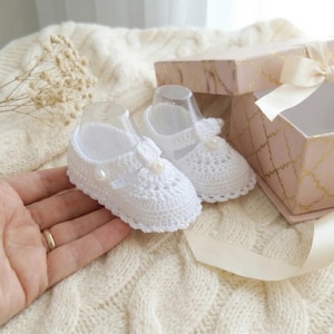 Puede incluir: Un par de zapatos de bebé blancos, de ganchillo, con detalles de perlas, expuestos cerca de una caja de regalo rosa con un patrón geométrico dorado y un lazo color crema. Los zapatos están sobre una superficie de punto color crema.
