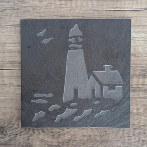 May include: A square, dark gray slate coaster featuring an etched design of a lighthouse, a small building, waves, and two birds in flight. The lighthouse is tall and conical, with a small building to its side.