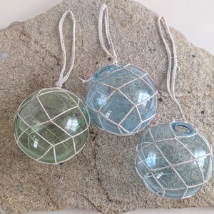 May include: Three glass fishing floats in shades of green and blue, encased in white netting, are displayed on a textured gray stone. Each float has a white string for hanging, suggesting a nautical or coastal theme.