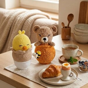 May include: A cozy breakfast scene featuring handmade crochet egg cups with a yellow chick and a brown bear. A croissant, a soft-boiled egg, and a cup of tea are also on the table. A small bowl of blueberries and granola complete the setting.