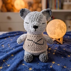 May include: A plush crochet animal toy featuring a light gray and white variegated yarn body with dark gray ear tips. The toy wears a beige sweater with a black embroidered constellation design on the front, closed eyes, and a small black nose.